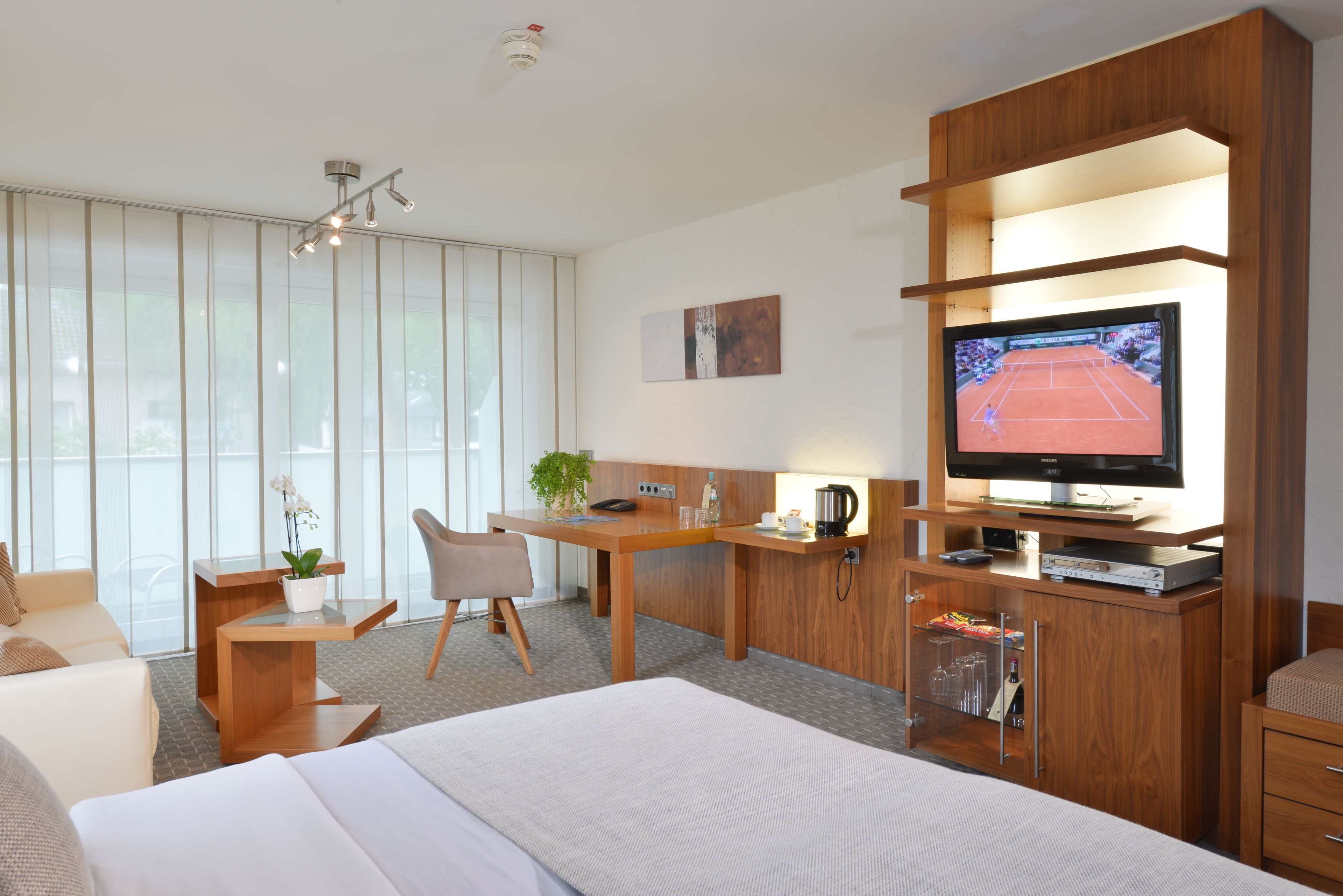 A modern hotel room with a large bed, a desk with a chair, a television, and a small cabinet.