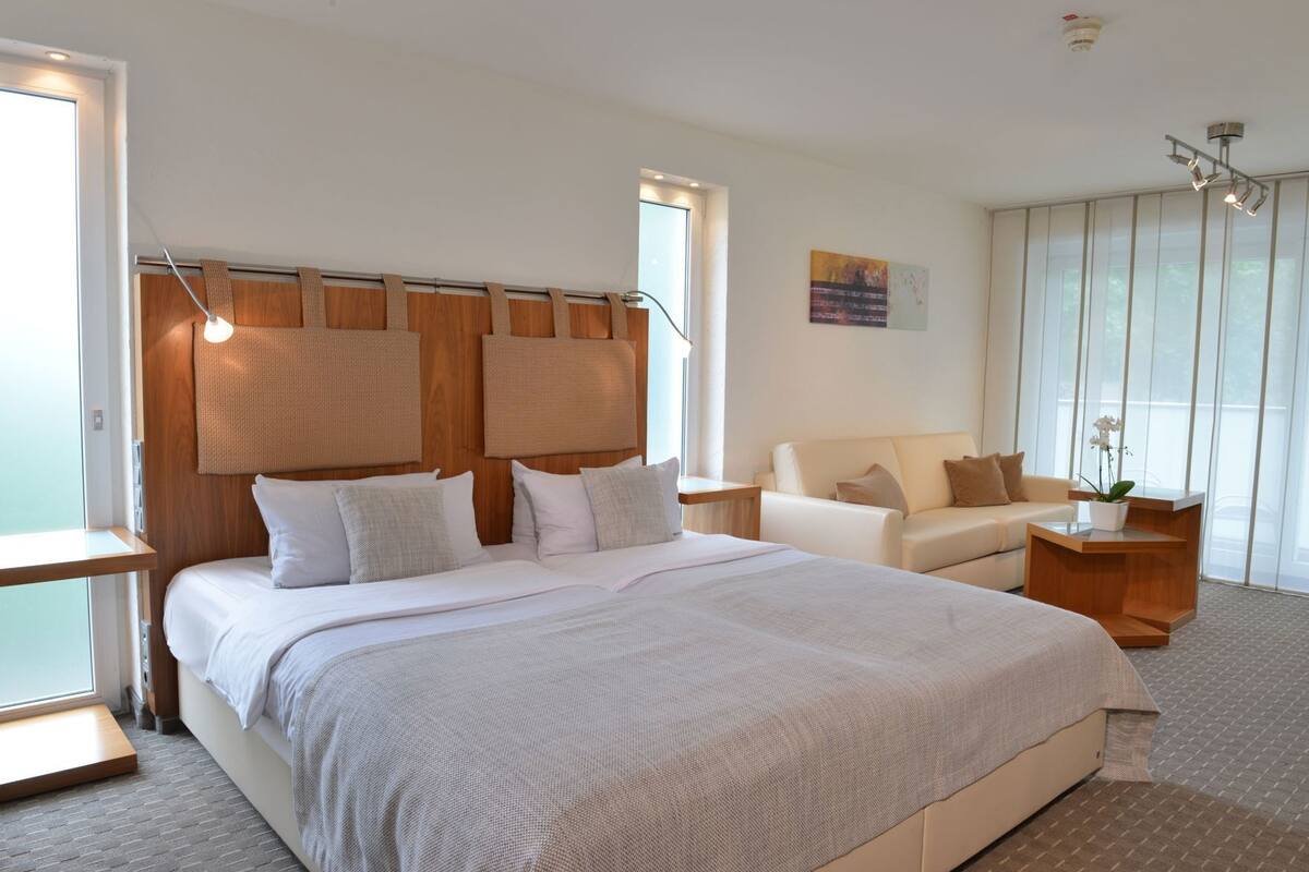 A modern hotel room with a large bed, a sofa, a small table, and a potted plant.