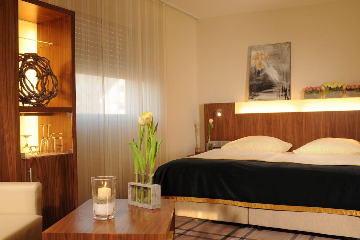A hotel room with a bed, a bedside table with a candle, a vase with flowers, a wall-mounted artwork, and a wooden shelf with glasses.