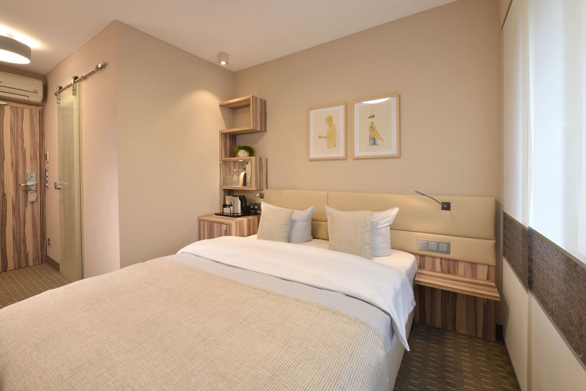 A hotel room with a bed, bedside tables, a wardrobe, a small shelf with items, and two framed pictures on the wall.
