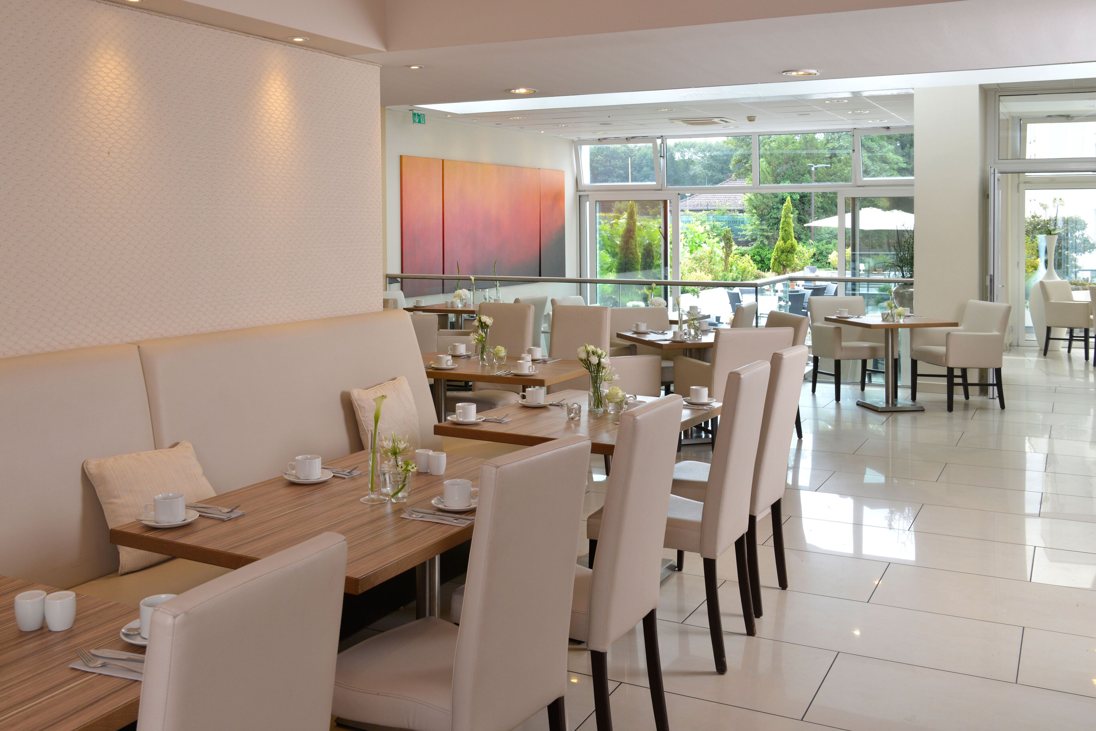 A modern dining area with multiple tables and chairs, large windows offering a view of greenery outside.