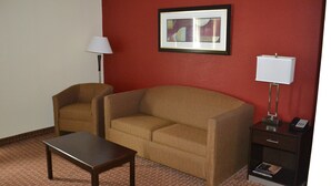 A room with a brown sofa, a small table, a lamp, a framed picture, and a nightstand with a phone and book. - Holiday Inn Express Harrisburg SW - Mechanicsburg by IHG (Mechanicsburg)