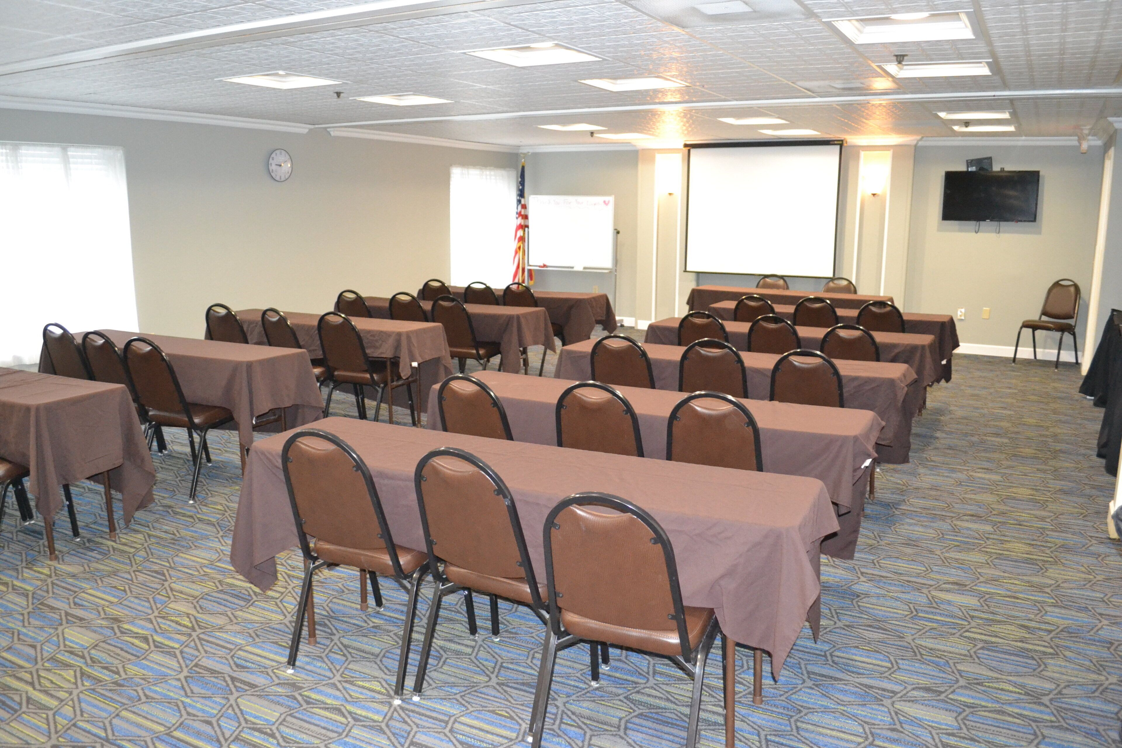 Uma sala de conferências com várias mesas e cadeiras, um relógio na parede e uma tela de projetor.