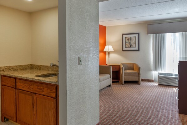 Habitación de hotel con lavamanos de encimera de granito, muebles de madera, una pequeña mesa con lámpara, una silla y un cuadro en la pared.
