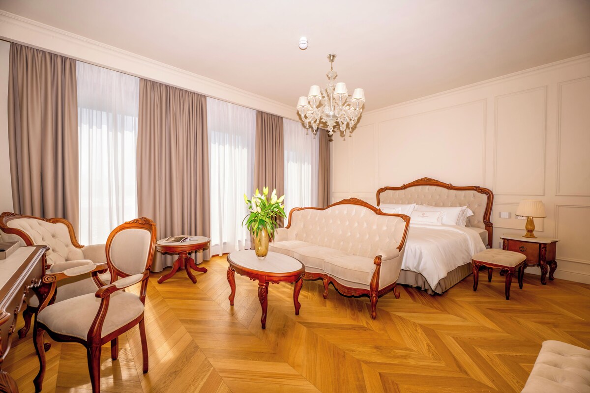 A hotel room with a large bed, a sitting area with a sofa and chairs, a small table with a vase of flowers, and a chandelier.