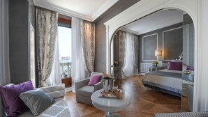 A spacious hotel room with a large bed, a sitting area with a sofa and armchairs, a round table, and a view of the city.