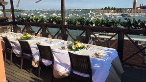 An outdoor dining area with tables set for two, overlooking a marina with boats and a prominent tower in the background.