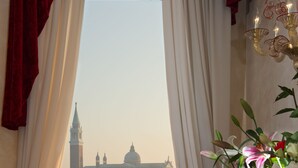 A room with a view of a historic building through a window, a table with a vase of flowers, and a chair with a red cushion.