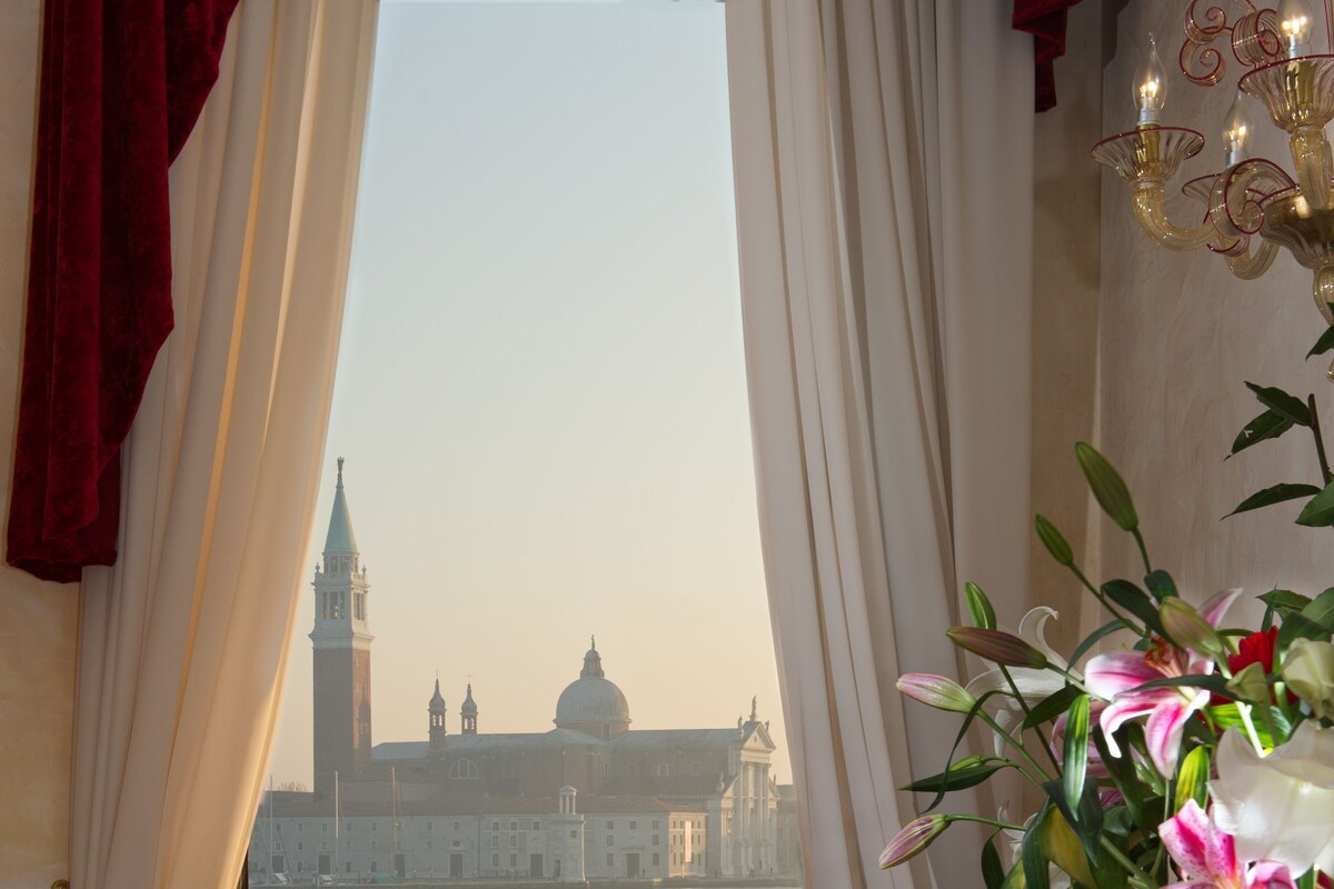 A room with a view of a historic building through a window, a table with a vase of flowers, and a chair with a red cushion.