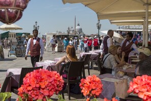 An outdoor café with tables and chairs, a street in the background, and a person walking. - Hotel Savoia e Jolanda, WorldHotels Elite (Venice)