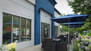 An outdoor patio area with tables and chairs, large windows, and a blue awning. - Hampton Inn Haverhill (Haverhill)