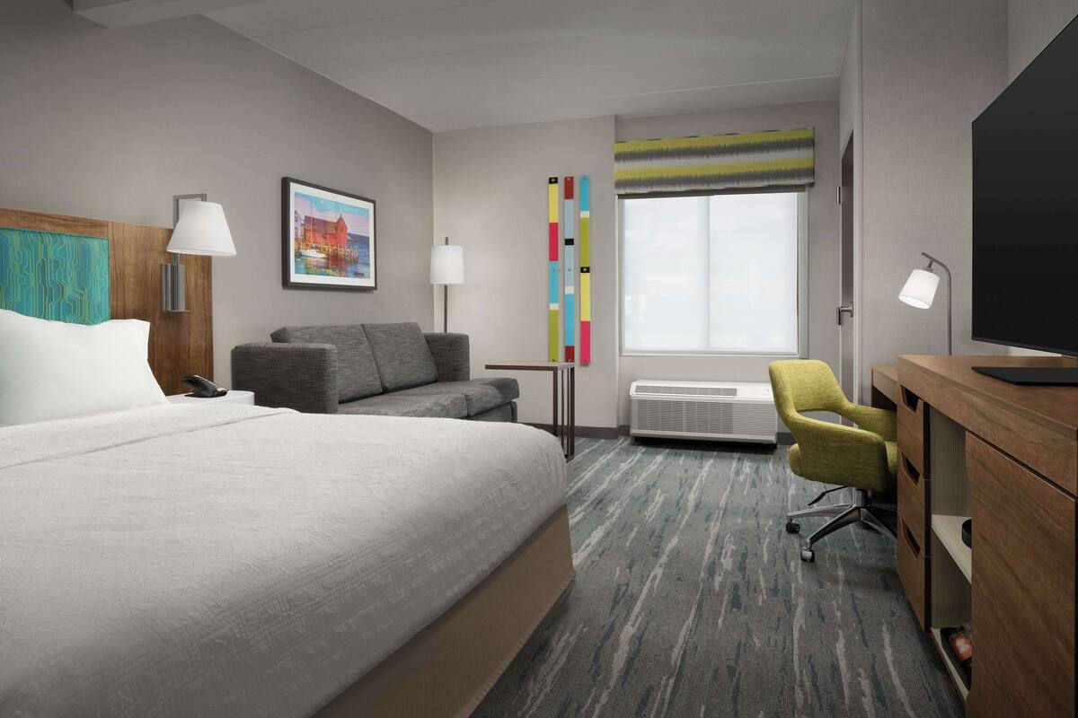 A hotel room with a large bed, a gray sofa, a desk with a chair, a flat-screen TV, and a window with a view.