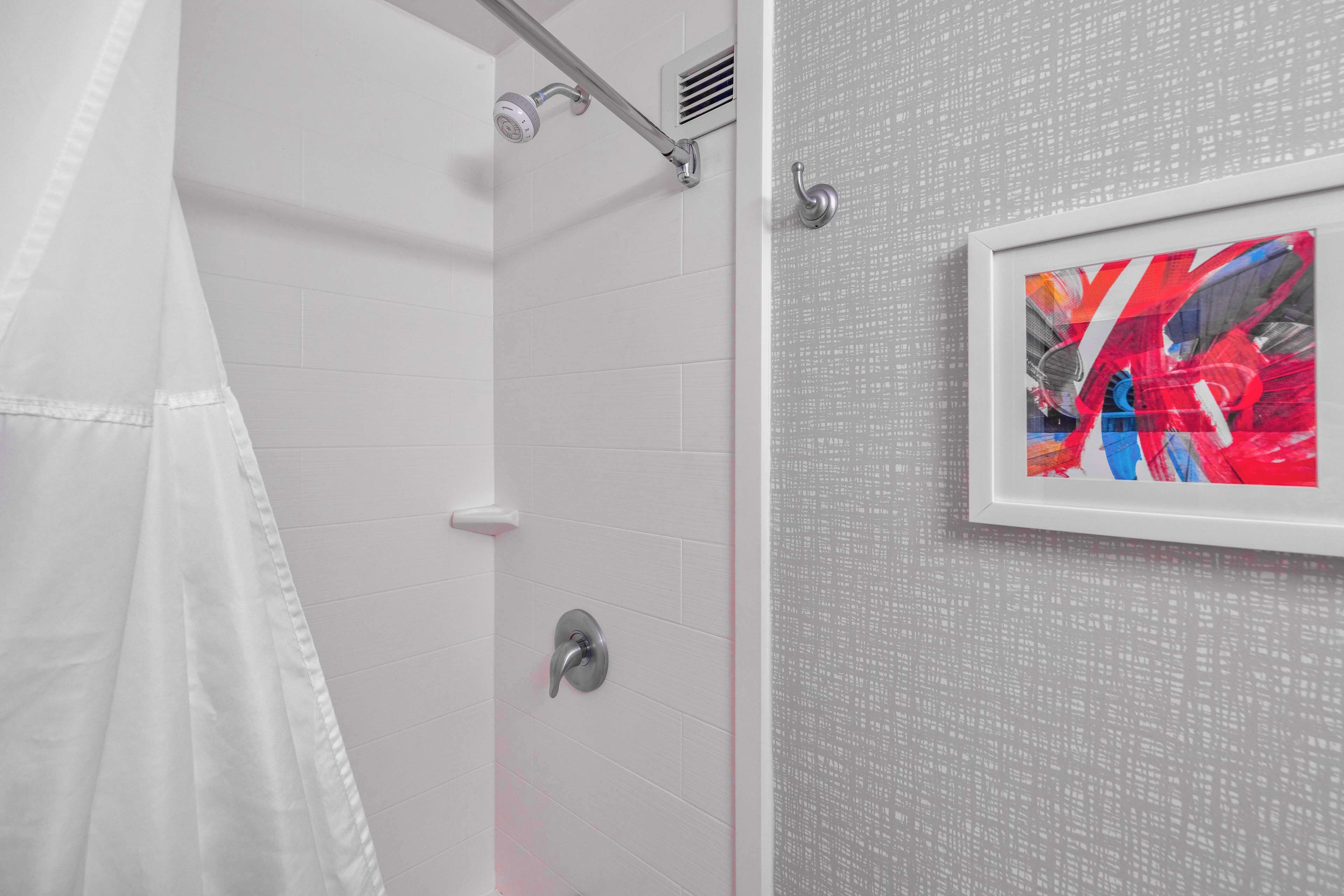 A bathroom with white tiles, a shower with a white curtain, and a framed abstract painting on the wall.