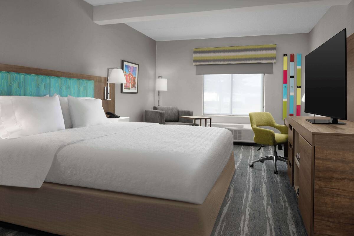 A hotel room with a large bed, a desk with a computer, a chair, a TV, and a window with a view.
