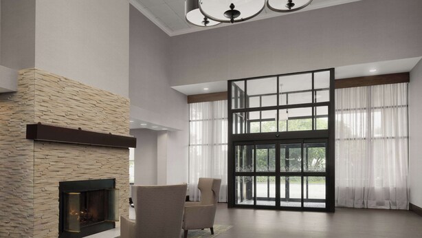 A spacious living room with a stone fireplace, large windows, and modern furniture. - Hampton Inn Haverhill (Haverhill)