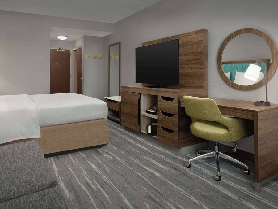 Quarto de hotel com cama, escrivaninha com televisão de tela plana, cadeira, espelho e piso coberto de carpete.