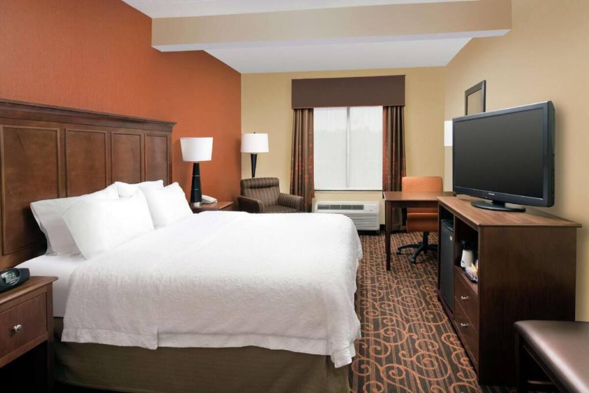 A hotel room with a large bed, a desk with a television, a chair, a lamp, and an air conditioning unit.