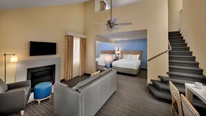 A hotel room with a flat-screen TV, a fireplace, a sofa, a bed, a desk, a lamp, a ceiling fan, and a staircase.
