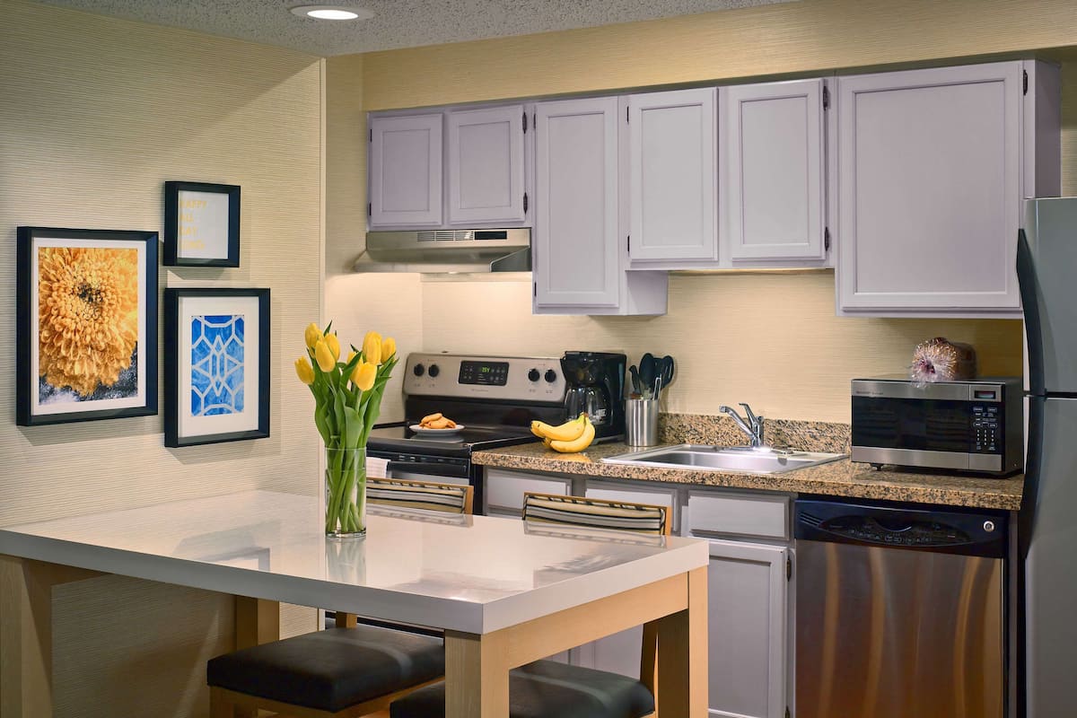A modern kitchen with white cabinets, a granite countertop, and stainless steel appliances. There is a dining table with two chairs and a vase of yellow flowers.
