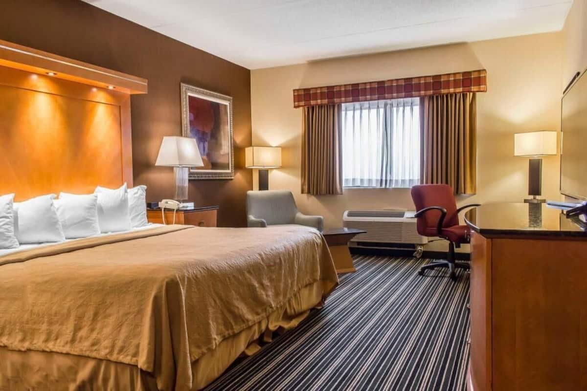 A hotel room with a large bed, a desk, a chair, a lamp, a window with curtains, and a painting on the wall.