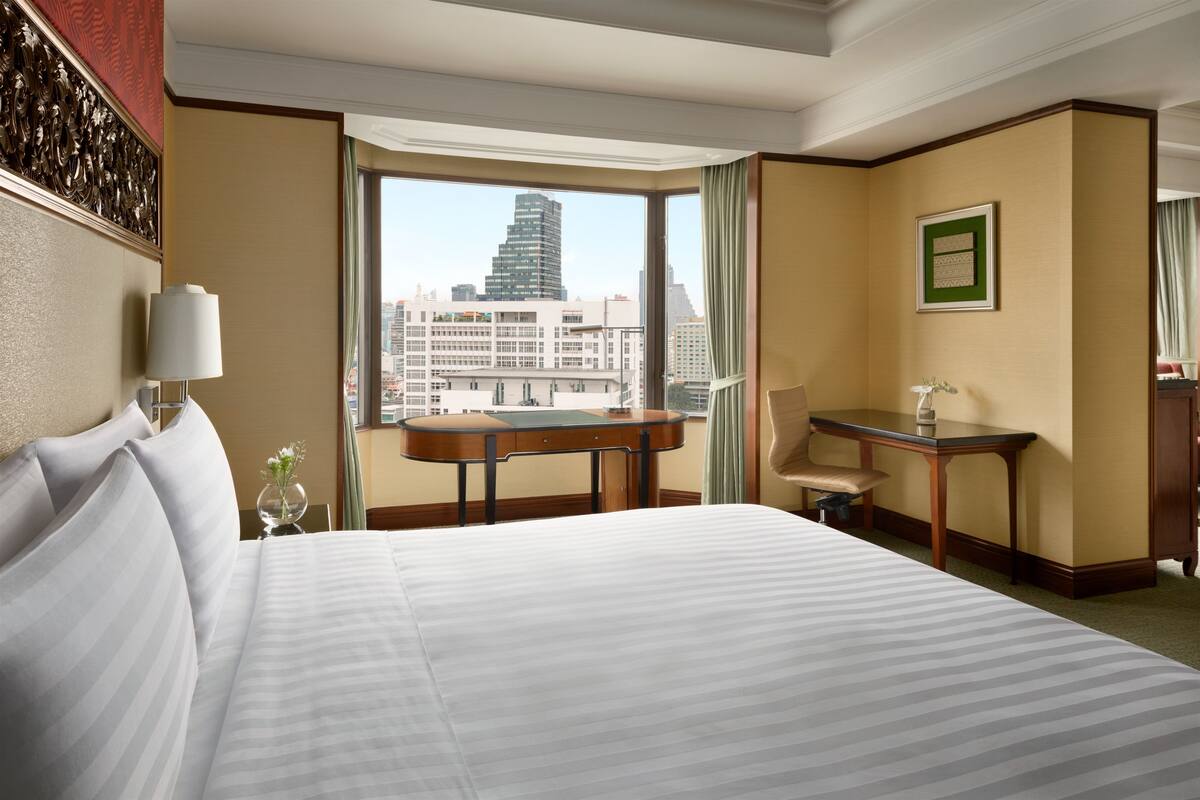A hotel room with a large bed, a desk, a chair, and a view of a cityscape through a large window.