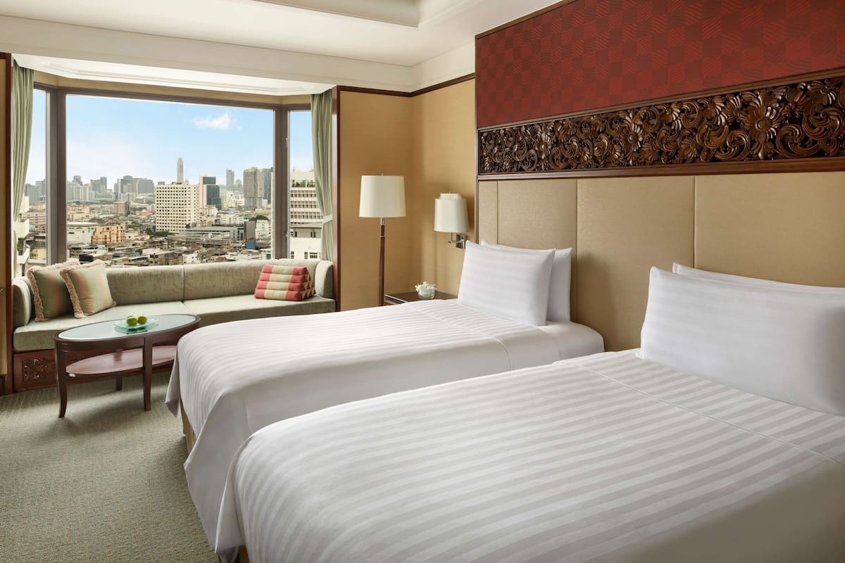 A hotel room with two beds, a sofa, a small table, and a view of a cityscape.