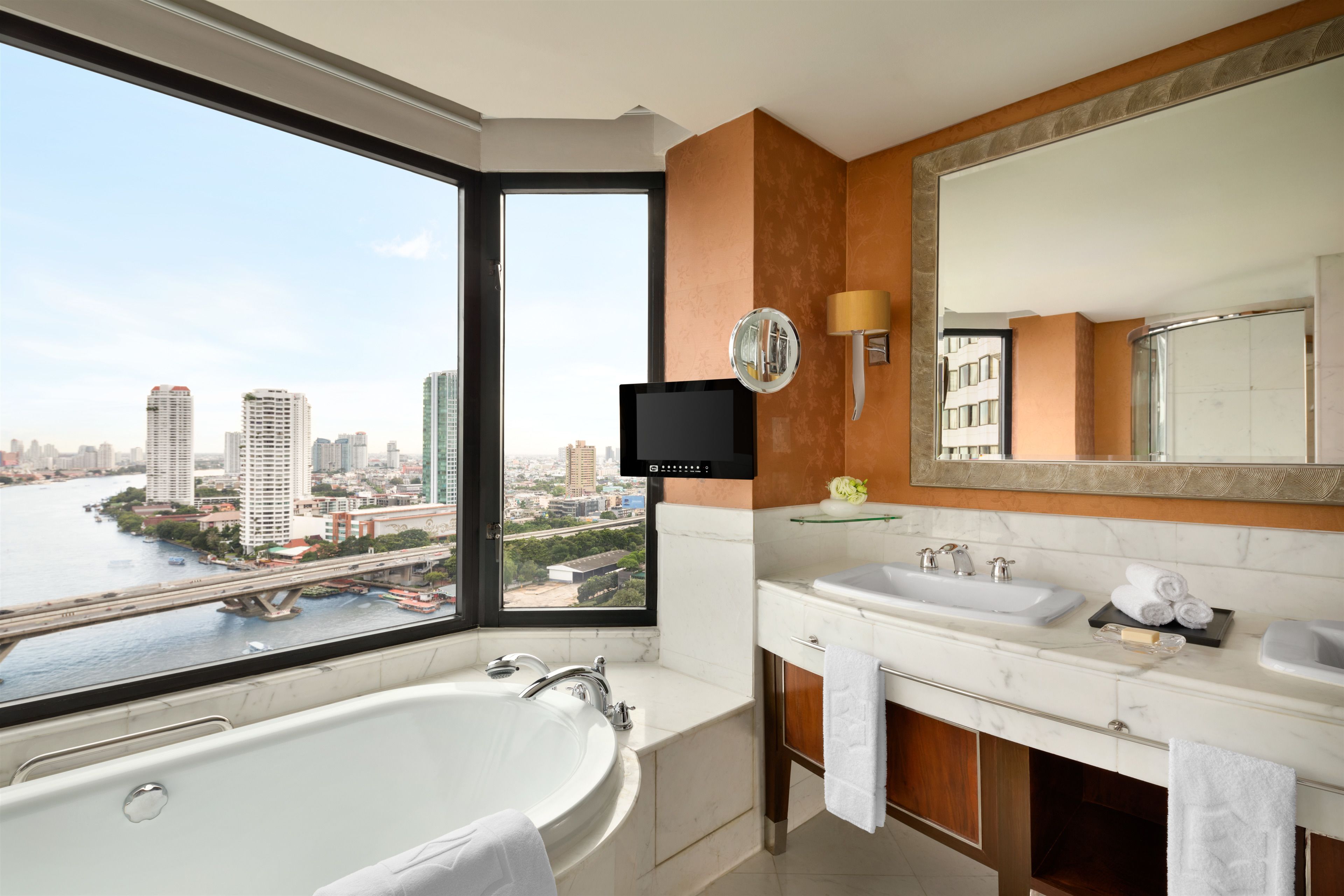 Un bagno moderno con una vasca da bagno grande, due lavandini e un ampio specchio. Una televisione è montata a parete e si apre una vista sulla città attraverso una grande finestra.