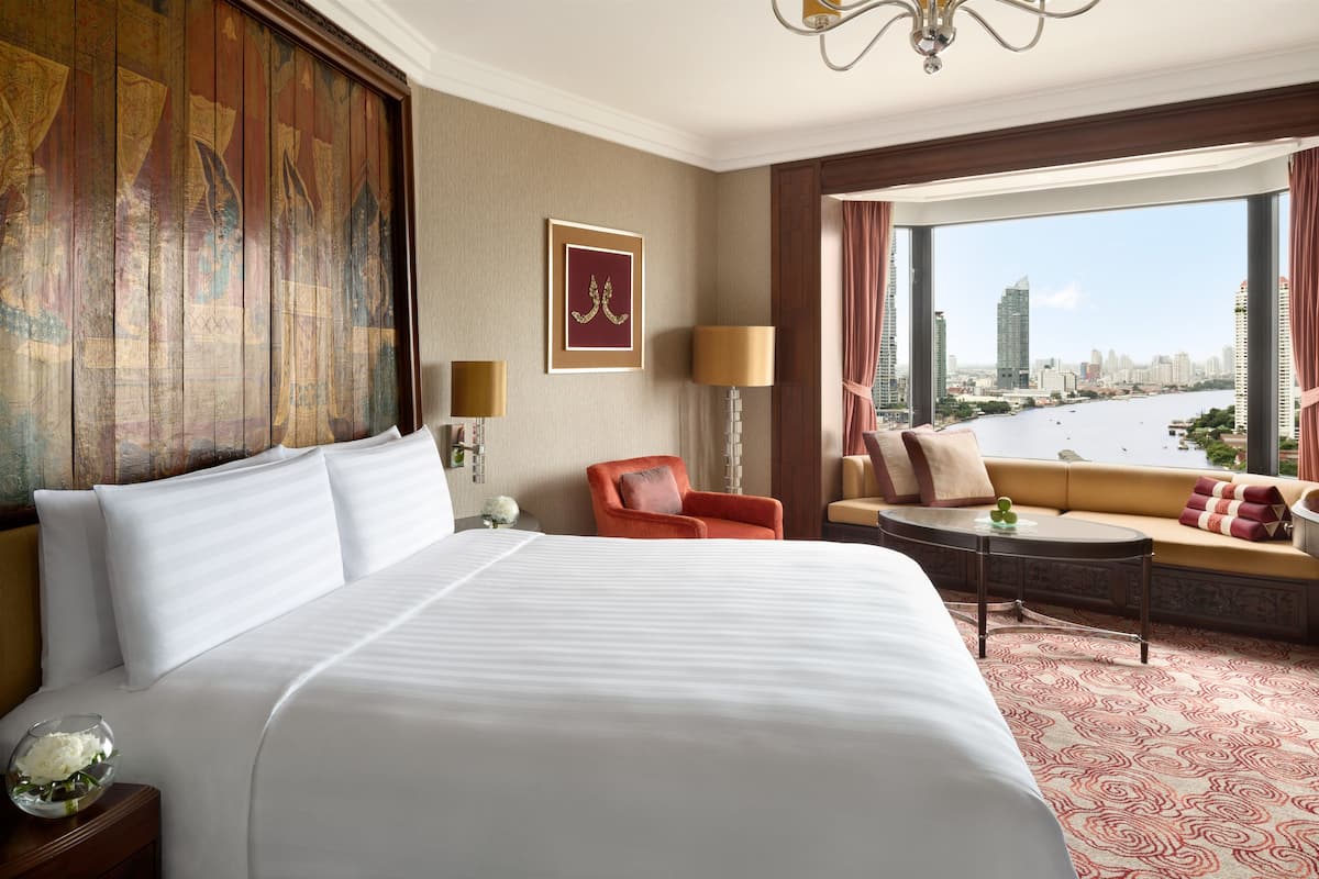 A hotel room with a large bed, a sofa, a small table, and a view of a cityscape through large windows.