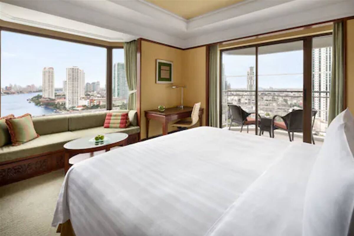 A hotel room with a large bed, a sofa, a desk, and a chair. There is a view of the city and a body of water through the windows.
