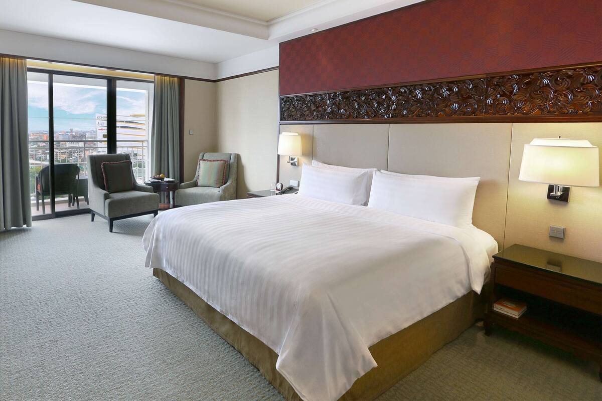 A hotel room with a large bed, two chairs, a desk, and a view of the city.
