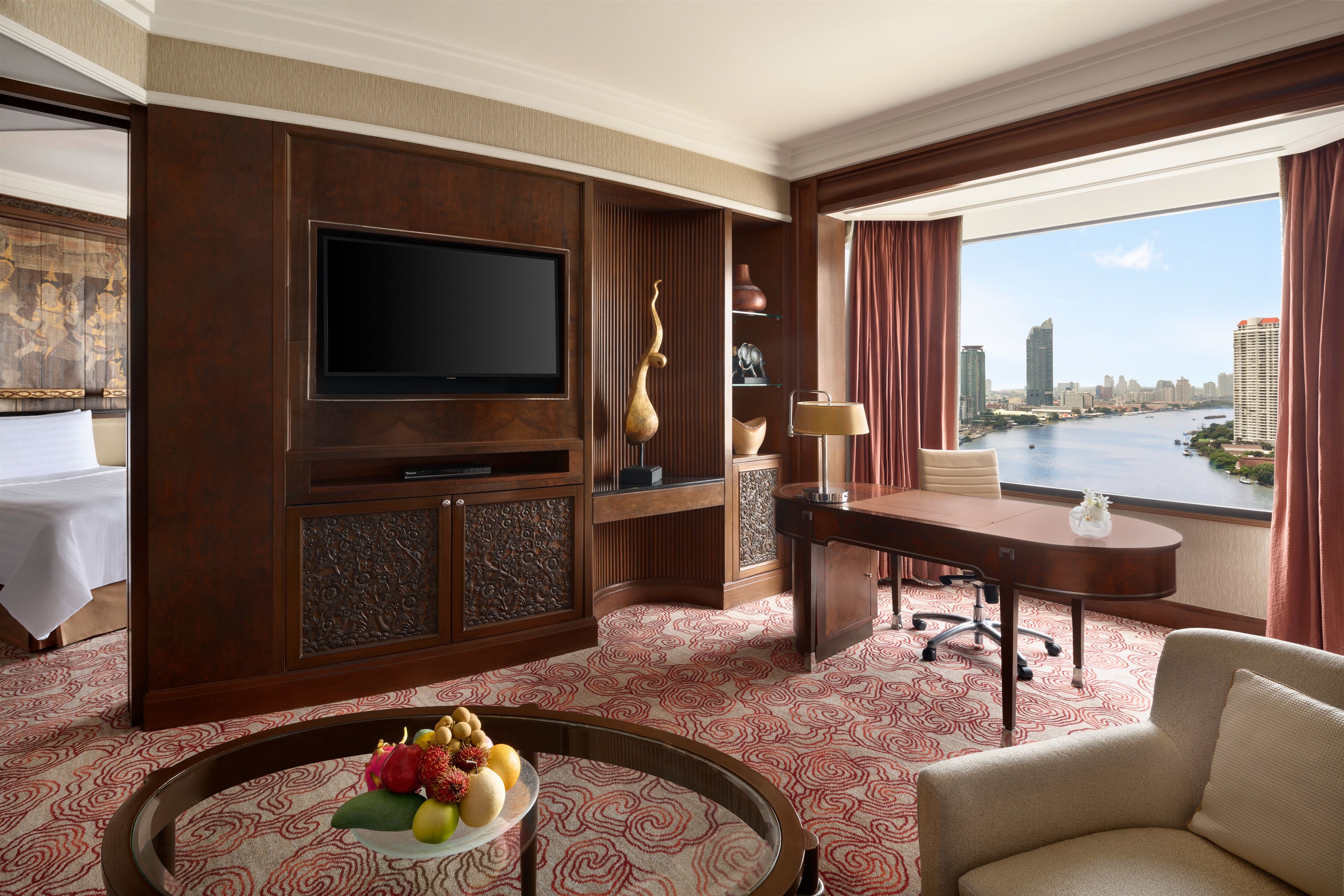 A hotel room with a large TV, a dining table with fruit, a desk with a lamp, and a view of a river and city skyline.