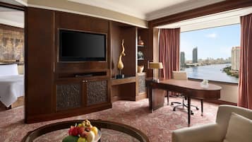 A hotel room with a large TV, a dining table with fruit, a desk with a lamp, and a view of a river and city skyline.