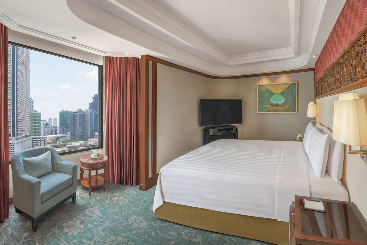 A hotel room with a large bed, a chair, a small table, a TV, and a view of the cityscape.