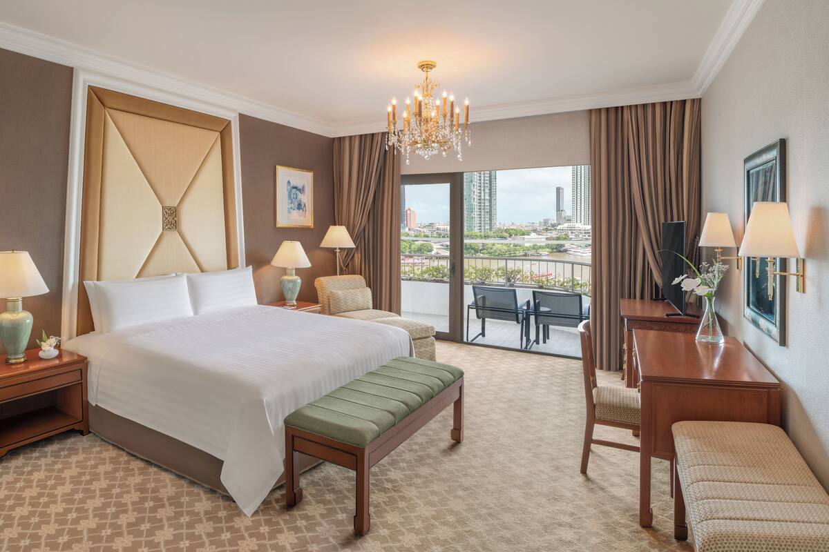 A hotel room with a large bed, a bench, a desk with a lamp, a chair, and a view of the city through the balcony.