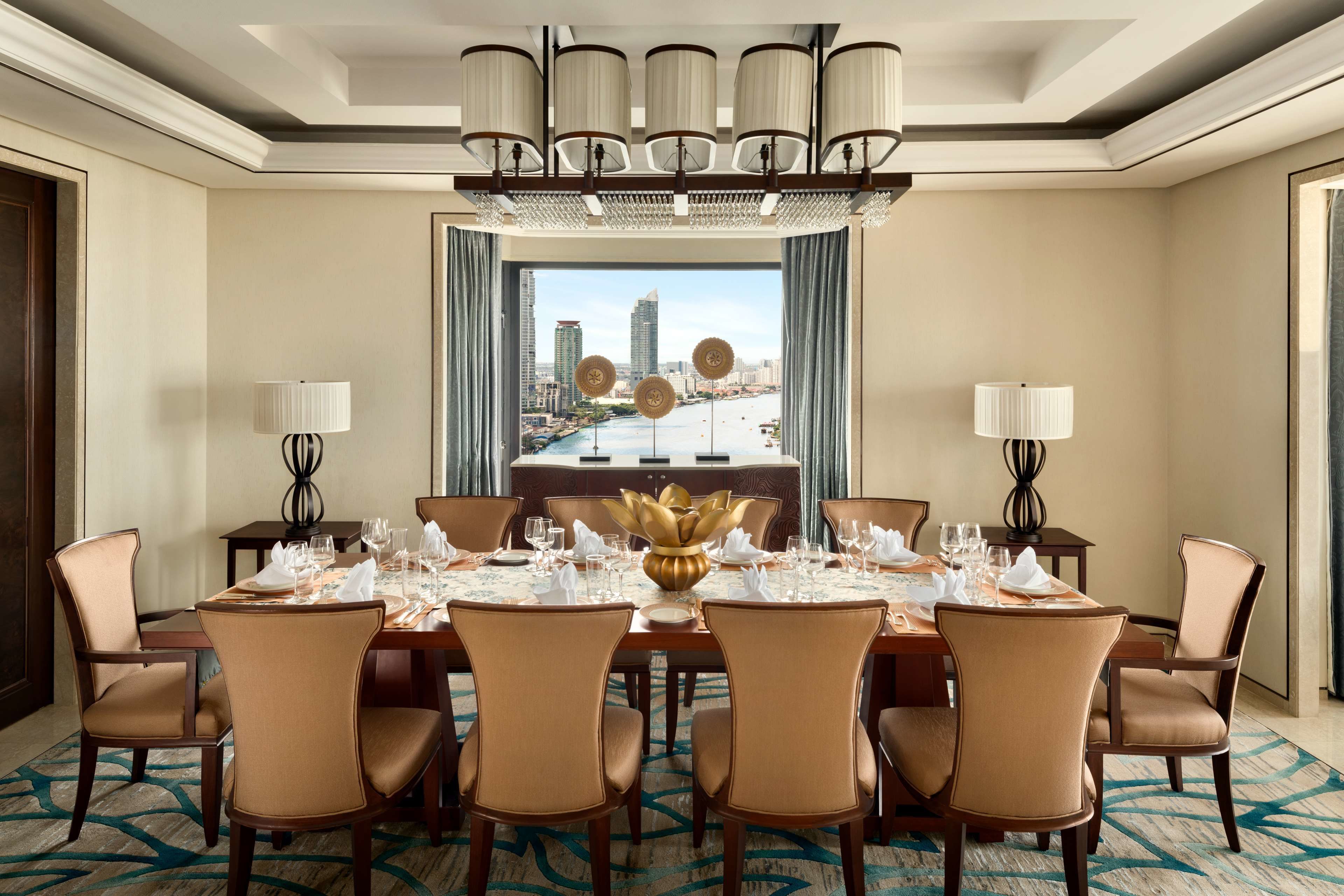 A hotel dining room with a large table set for six, surrounded by six chairs, a chandelier, and a view of a cityscape through large windows.
