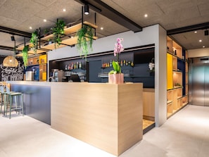 A modern bar area with a wooden counter, hanging plants, and shelves with bottles and glasses. - Ibis Paris Meudon Velizy (Meudon)