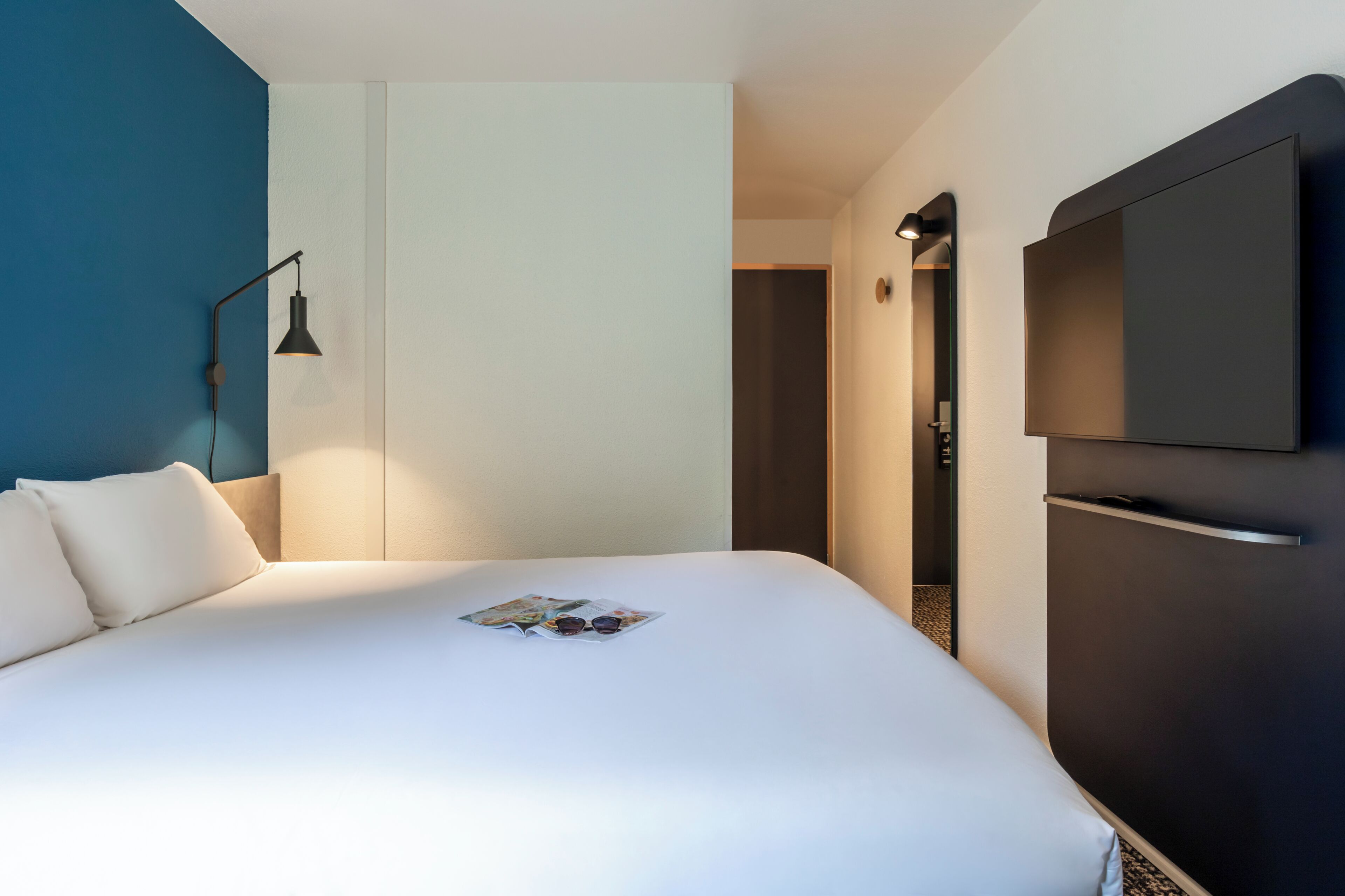 Een hotelkamer met een bed, een aan de muur bevestigde televisie en een deur naar een andere kamer.