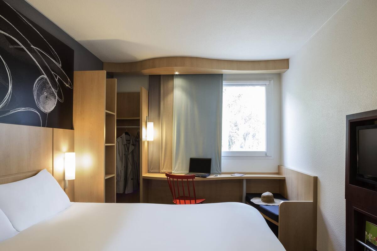 A hotel room with a bed, a desk with a laptop, a chair, a TV, and a wardrobe.