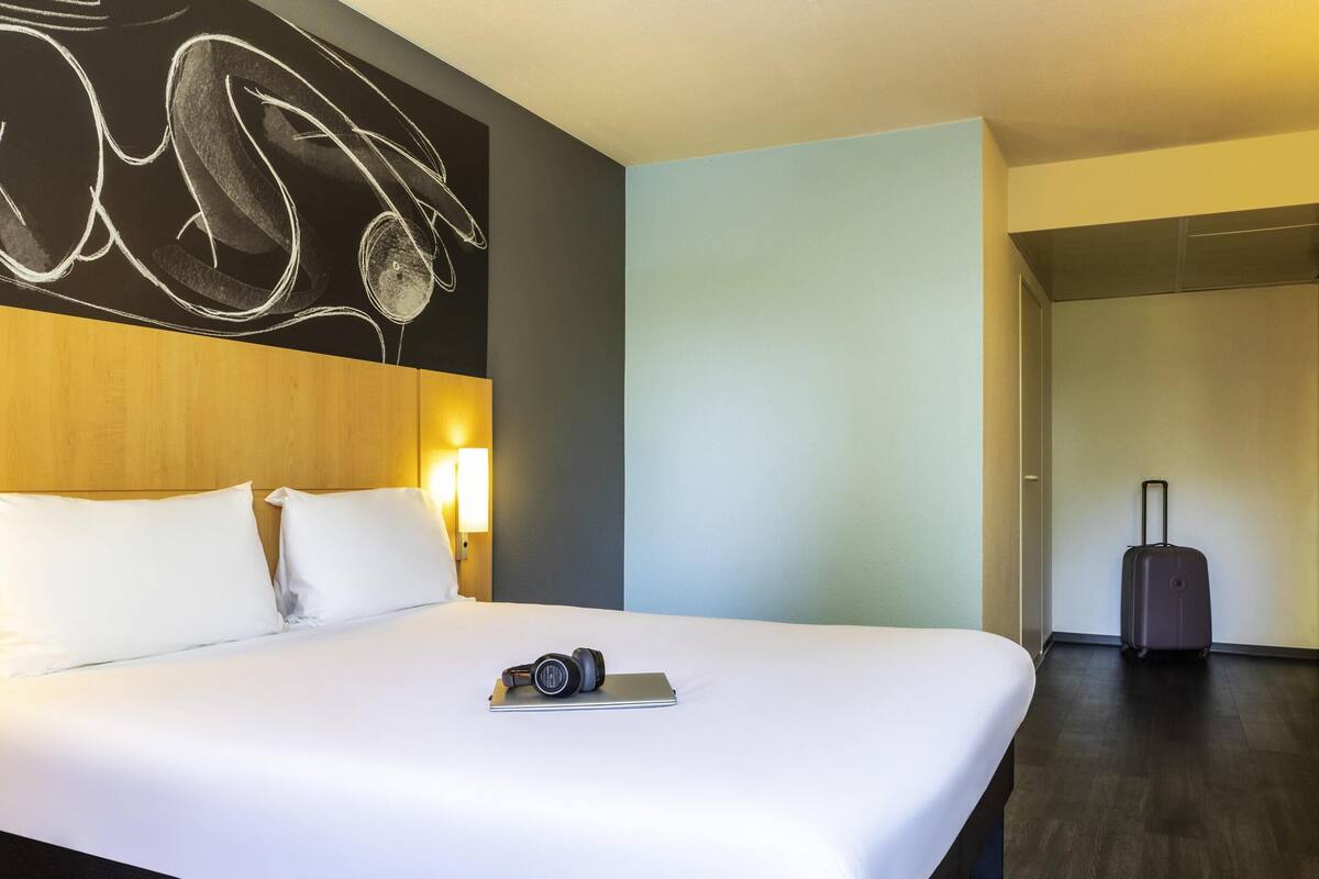A hotel room with a bed, a headboard, a wall art piece, a lamp, and a suitcase.