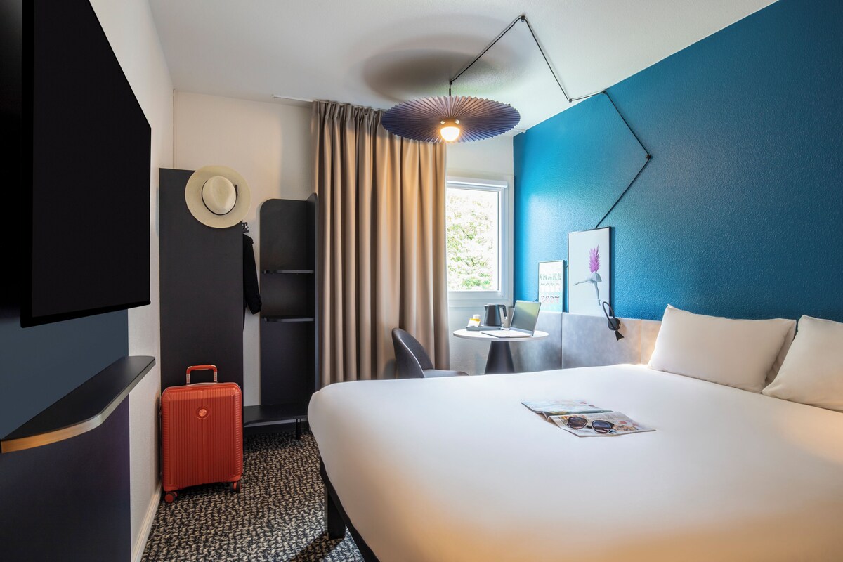 A hotel room with a bed, a desk with a laptop, a suitcase, a TV, and a window with curtains.