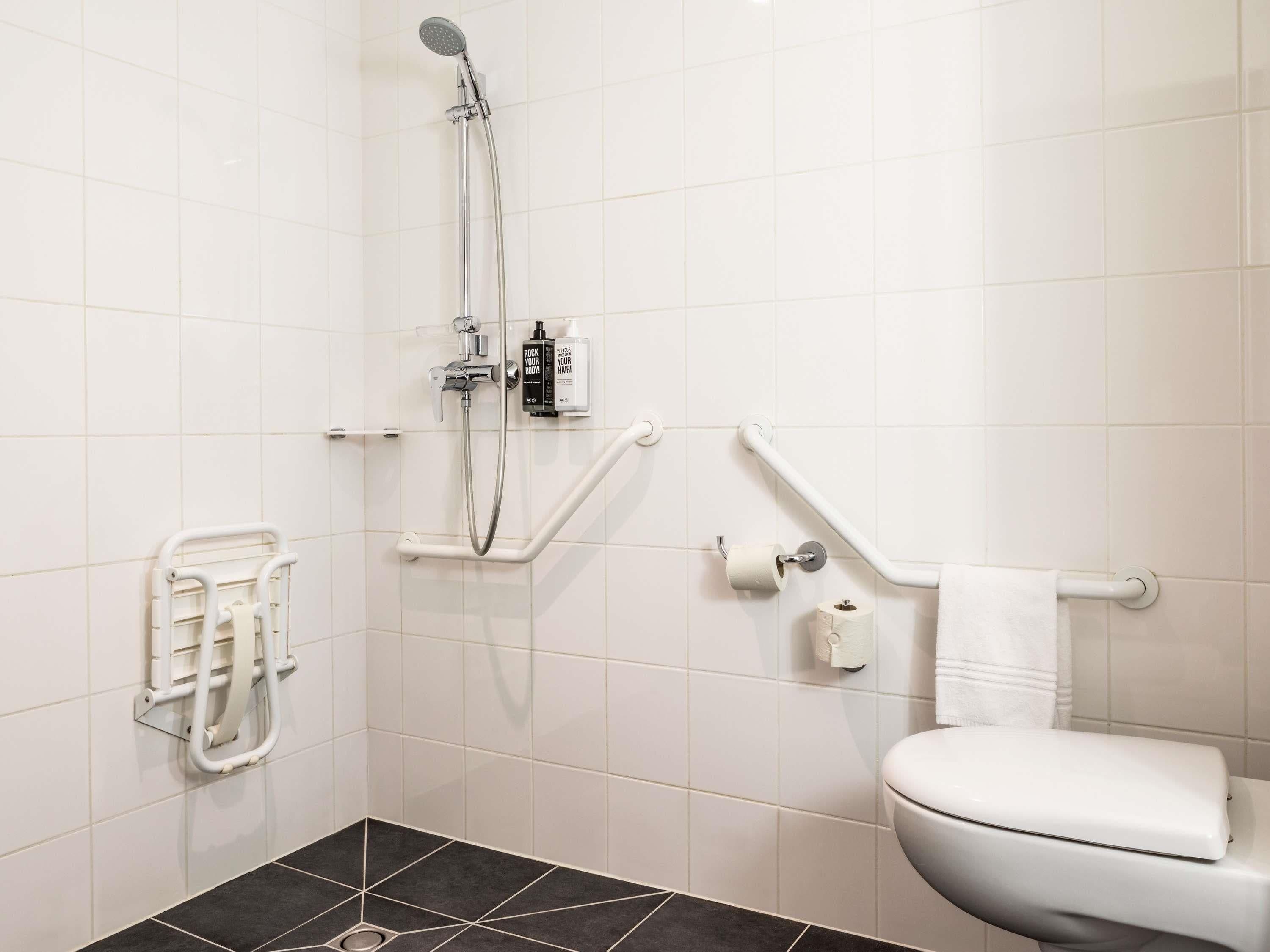 A bathroom with a white toilet, a shower with a handheld showerhead, grab bars, and a roll of toilet paper on the wall.