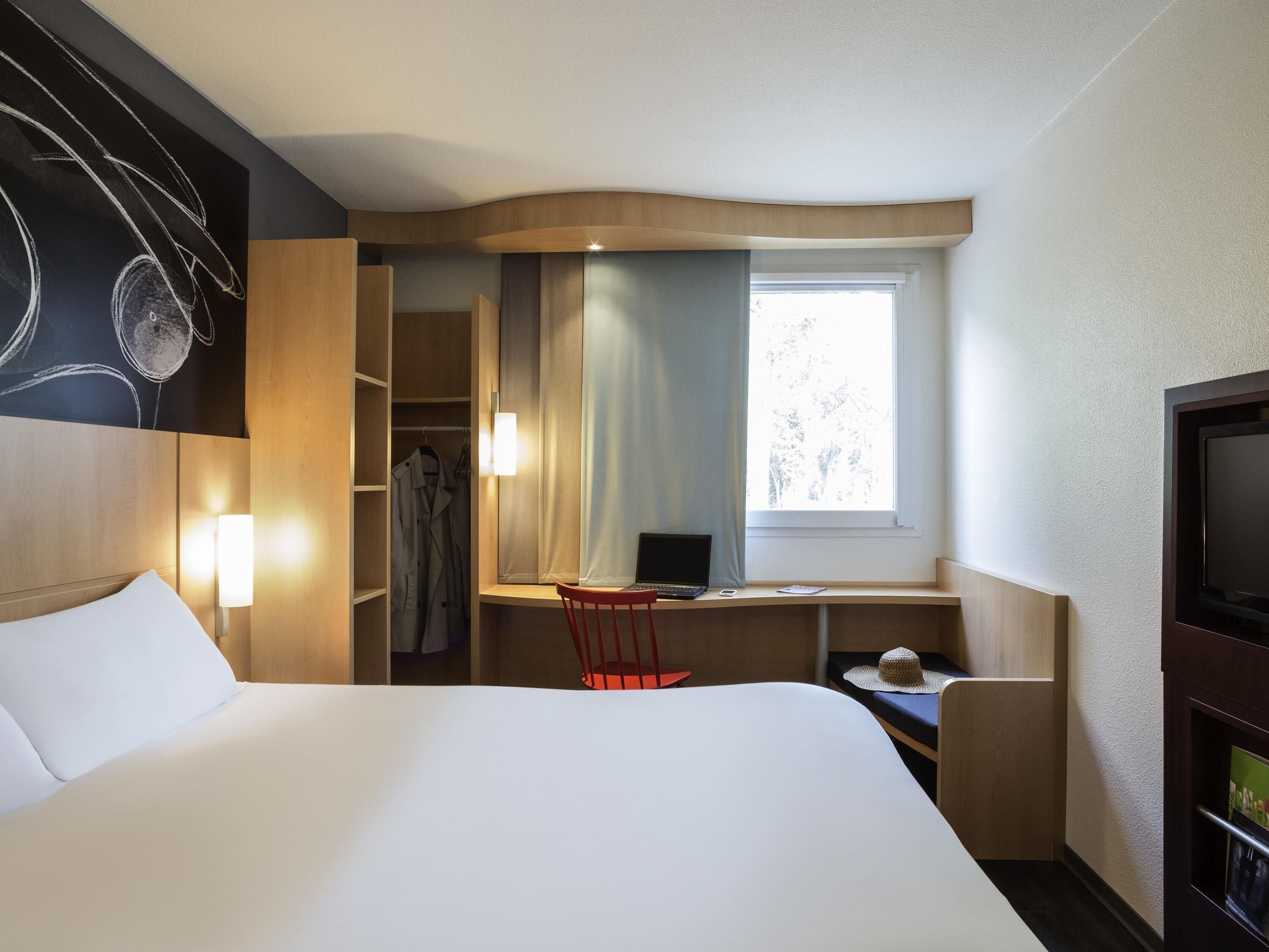 A hotel room with a bed, a desk with a laptop, a chair, a TV, and a wardrobe.