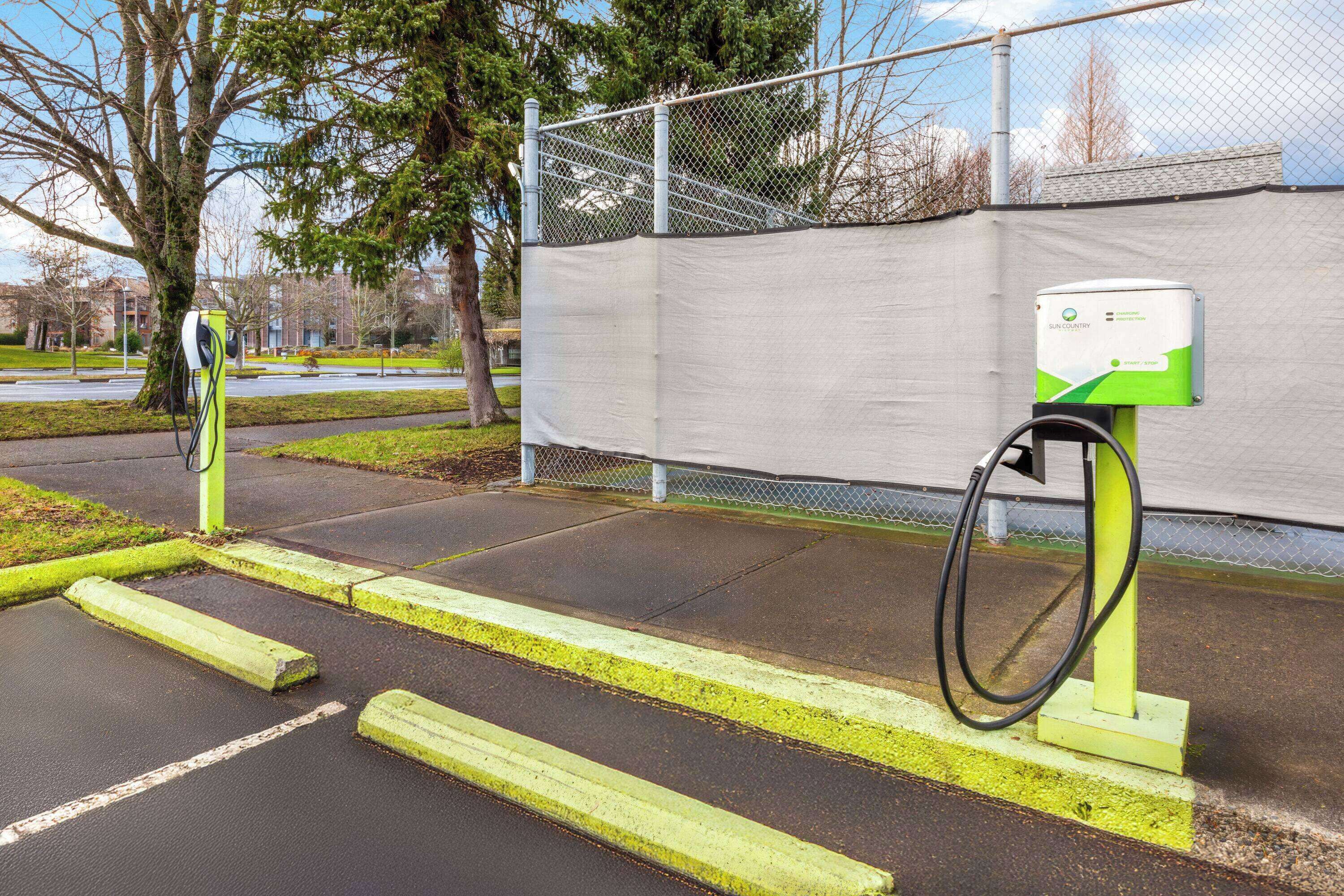 Una stazione di ricarica per veicoli elettrici con un box verde e bianco, un cavo di ricarica e cordoli gialli.