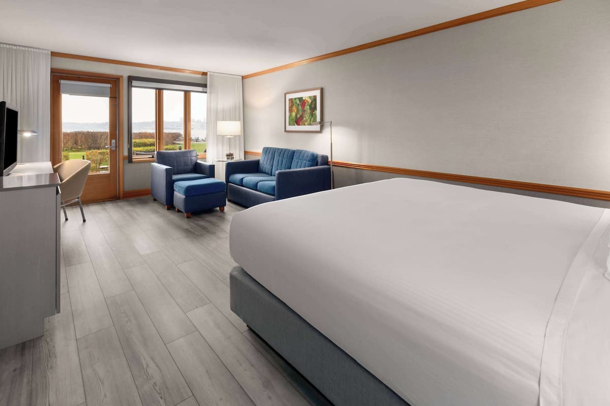 A hotel room with a large bed, a desk with a chair, two armchairs, a TV, and a window with a view of the outdoors.