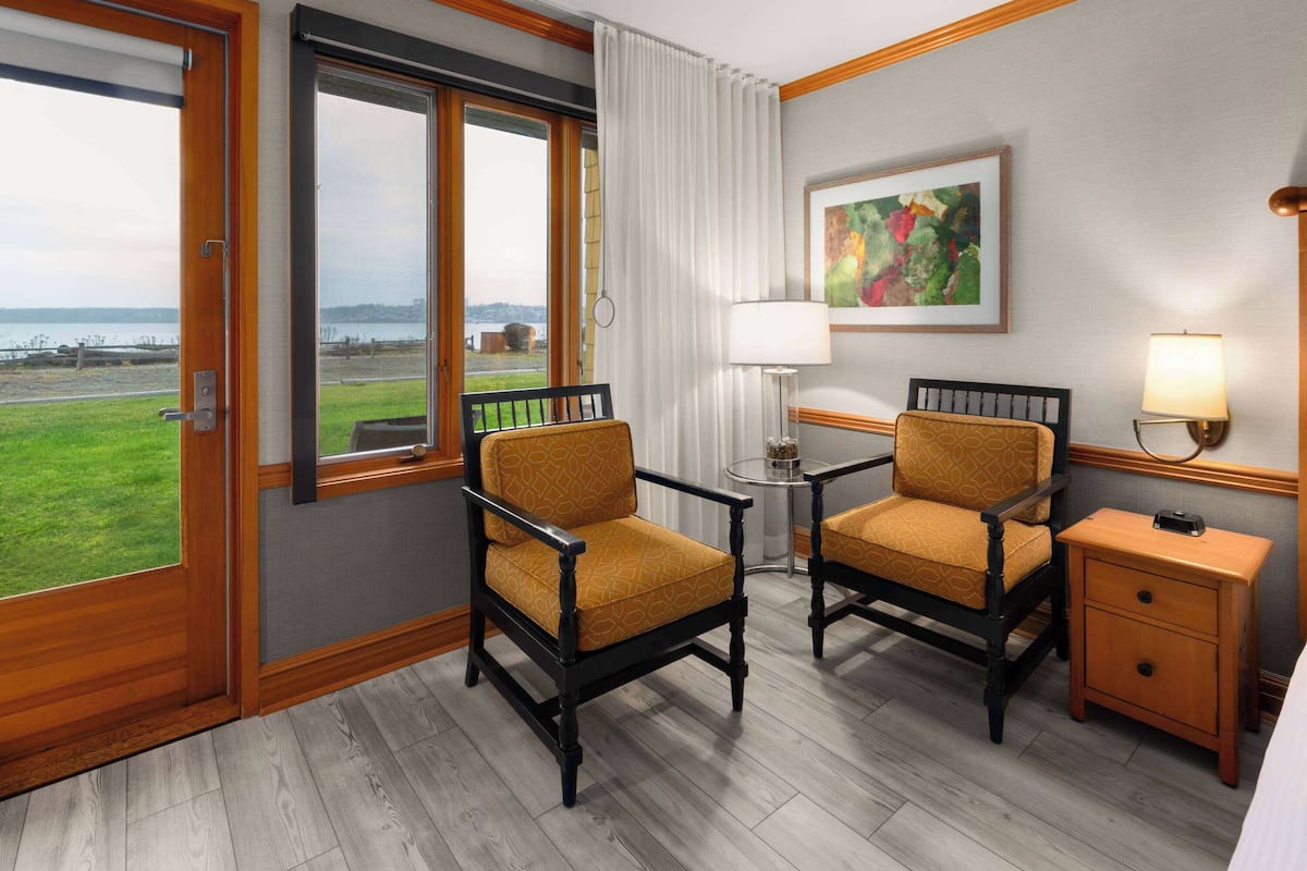 A hotel room with two armchairs, a small table, a bedside table, a lamp, and a painting on the wall.