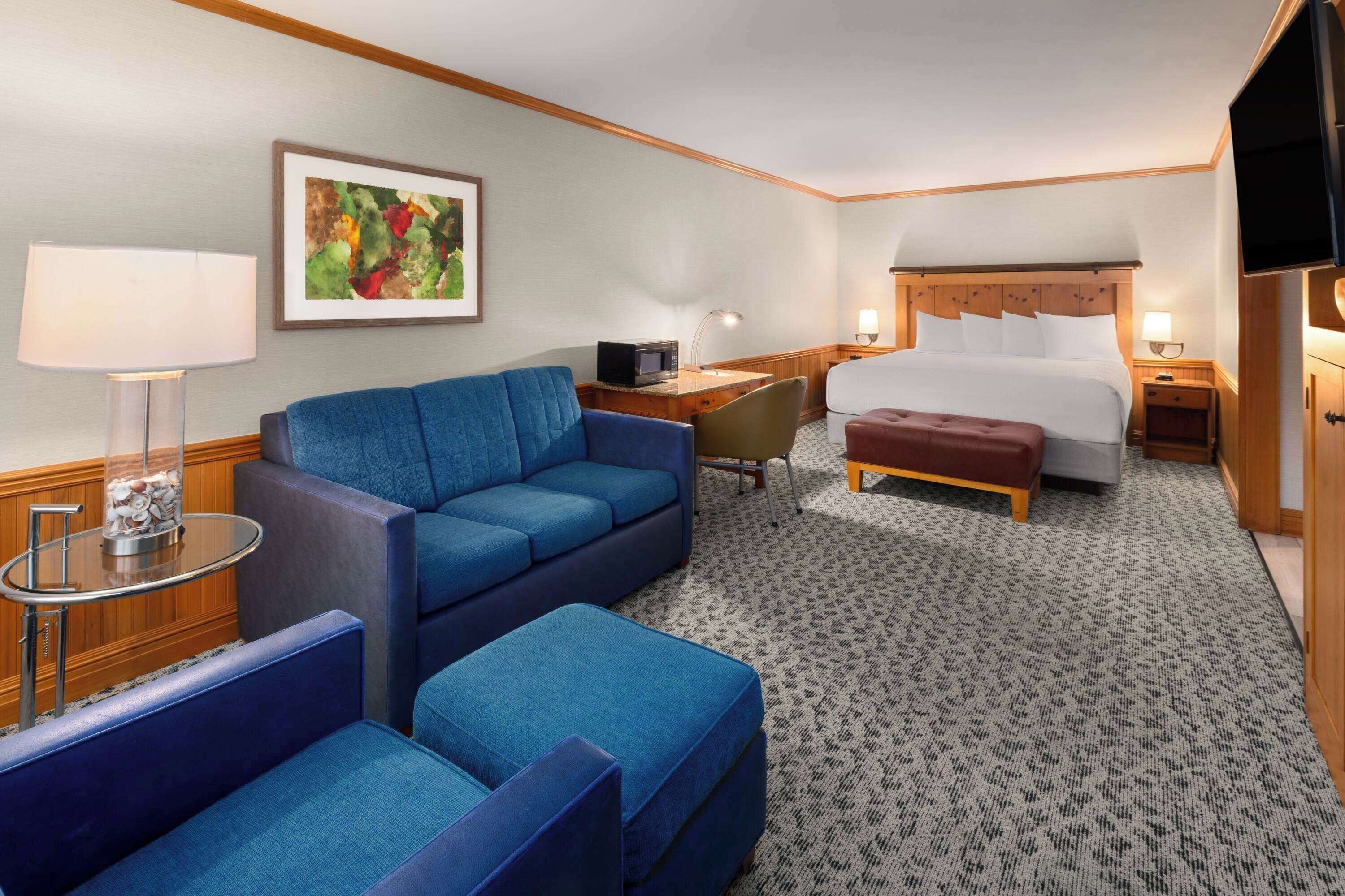 A hotel room with a bed, a blue sofa, a desk with a lamp, and a television.