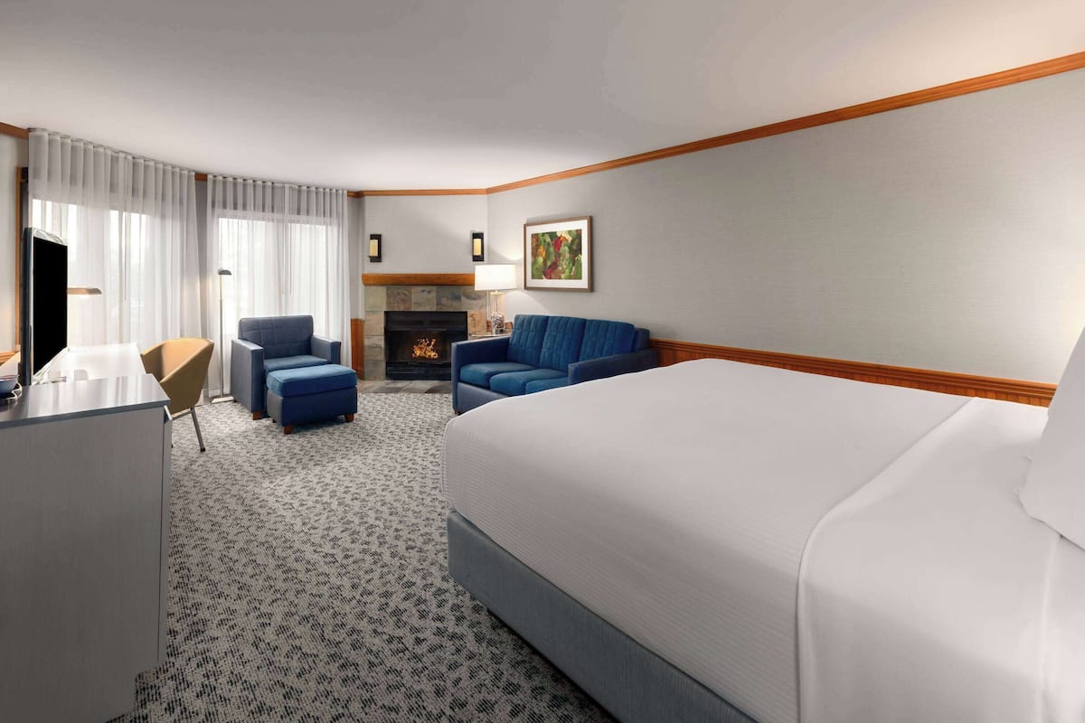 A hotel room with a bed, two blue armchairs, a desk, a fireplace, and a framed picture on the wall.