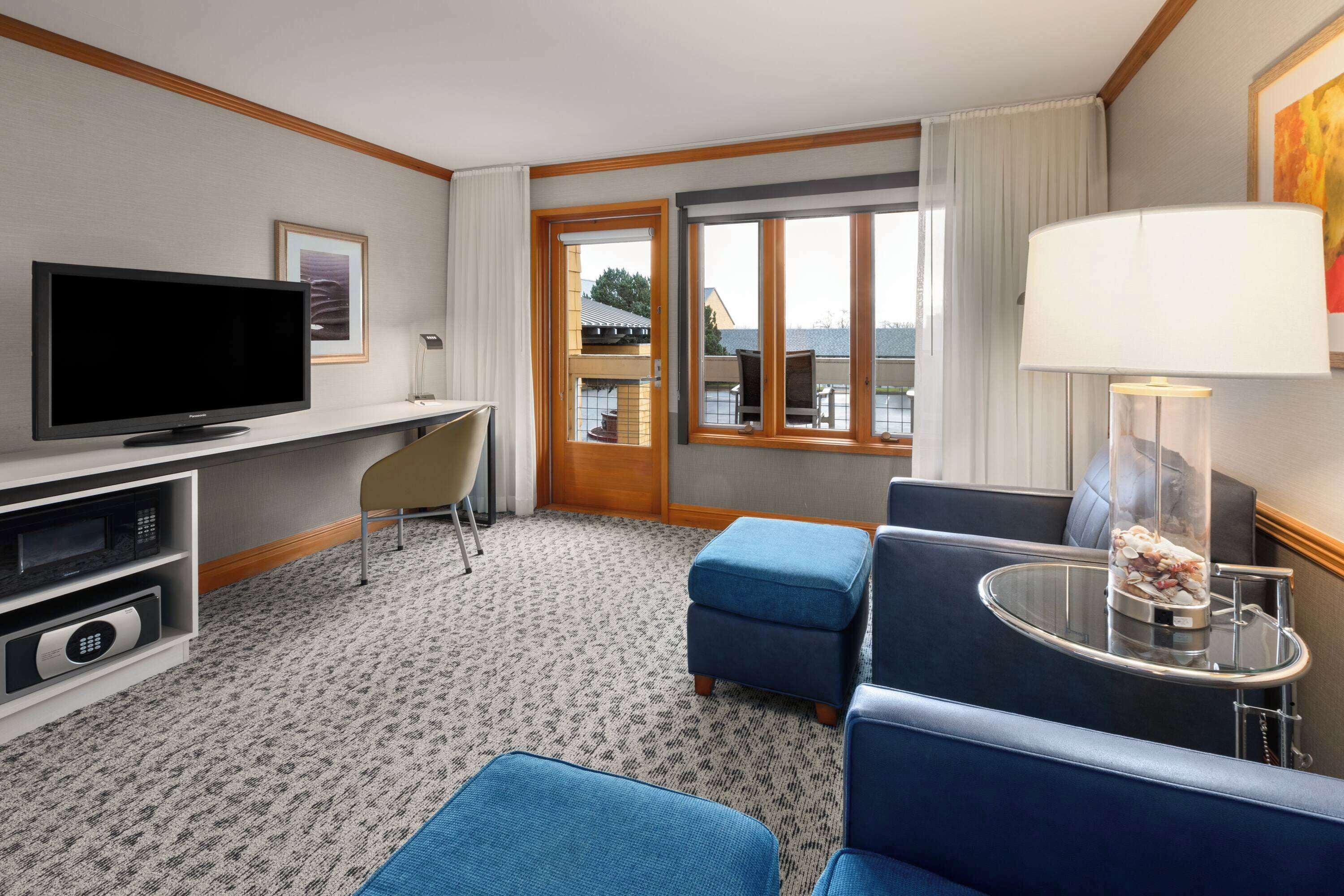Hotelkamer met een flatscreen televisie, een bureau, een stoel, een bank, een klein tafeltje met een lamp, en uitzicht op de omgeving via een schuifdeur.