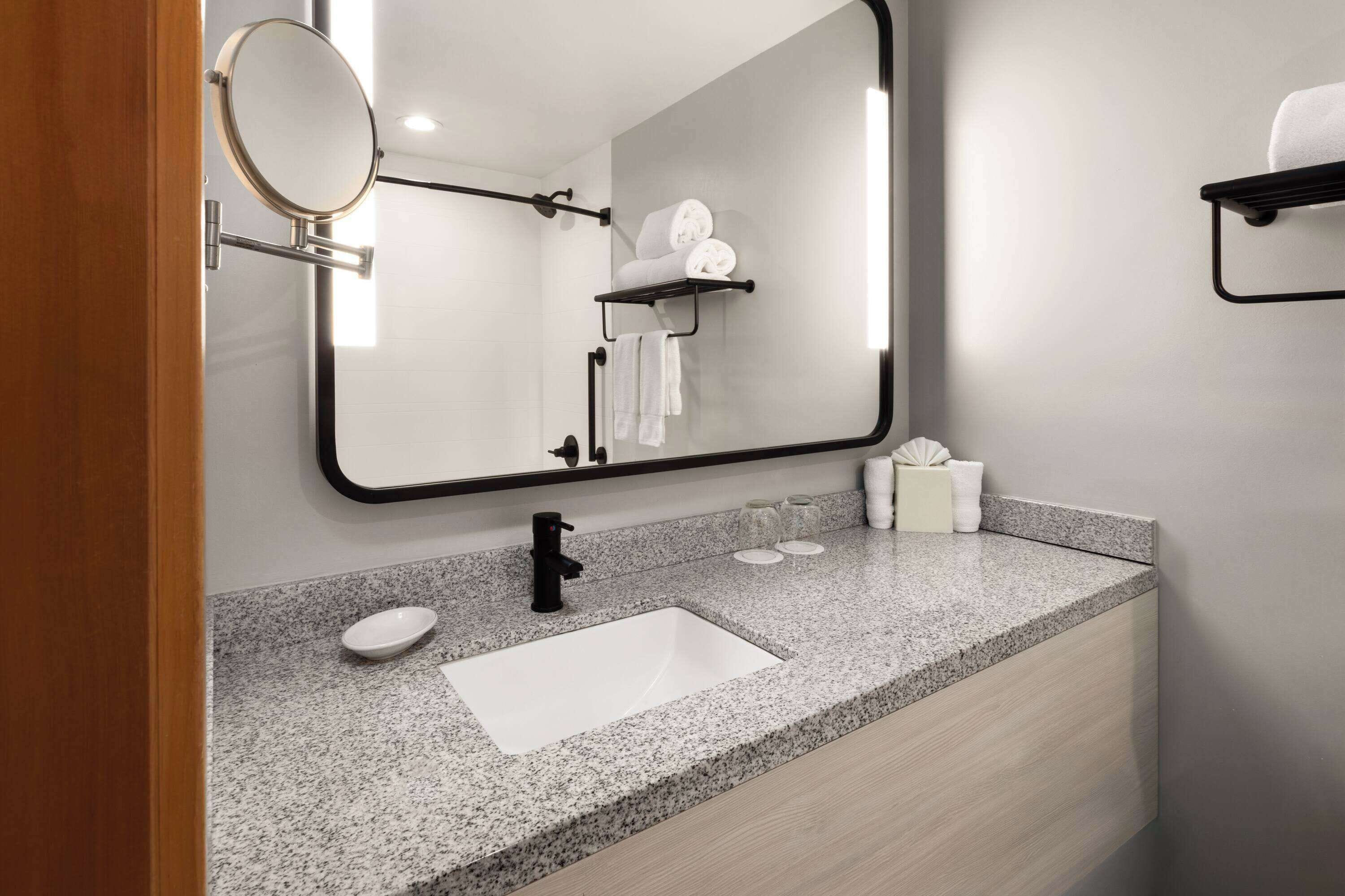 A modern bathroom with a granite countertop, a white sink, a black faucet, a round mirror, and a towel rack with folded towels.