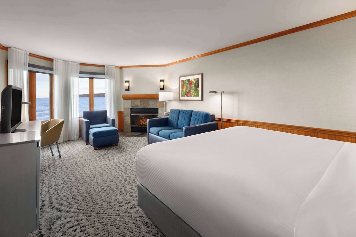 A hotel room with a large bed, a desk with a television, a seating area with a sofa and chairs, a fireplace, and a view of the sea.